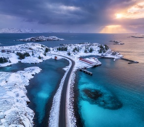 Aerial view of road, island in snow, rorbu, sea, bridge, snowy mountains, azure water, cloudy sky at sunset in winter. Dramatic landscape. Top drone view of road. Henningsvaer, Lofoten islands, Norway