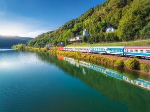 Aerial view of modern high speed train moving near river in alpine mountains at sunny sumer day. Top view of intercity train, railroad, lake, road, green trees in spring. Railway station in Slovenia