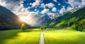 Aerial view of road in alpine mountains, green meadows, trees at sunset in summer. Top view of country road. Colorful landscape with road, rocks, field, grass, blue sky, clouds. Logar valley, Slovenia