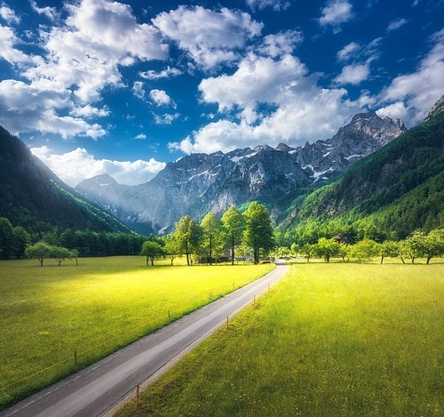 Aerial view of road in alpine mountains, green meadows, trees in summer. Top view of country road. Colorful landscape with road, rocks, field, grass, blue sky, sunbeam, clouds. Logar valley, Slovenia