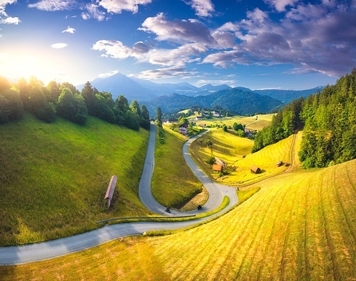 Aerial view of country road in green meadows at sunset in summer. Top drone view of rural road, alpine mountains. Colorful landscape with curved highway, hills, fields, green grass, blue sky. Slovenia