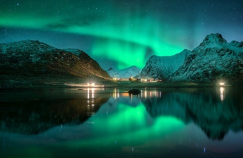 Aurora borealis, snowy mountains, sea, fjord, reflection in water, street lights at starry winter night. Lofoten, Norway. Northern lights. Landscape with polar lights, snowy rocks, sky with stars