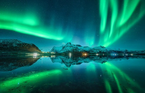 Aurora borealis, snowy mountains, sea, fjord, reflection in water, street lights at starry winter night. Lofoten, Norway. Northern lights. Landscape with polar lights, snowy rocks, sky with stars