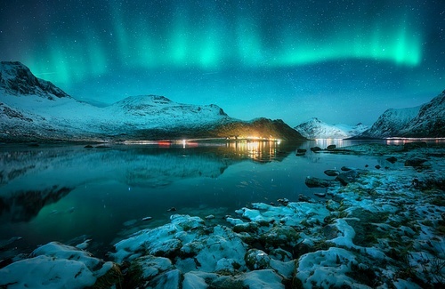 Aurora borealis over the snowy mountains, frozen sea, reflection in water at winter night in Lofoten, Norway. Northern lights and snowy rocks. Landscape with polar lights, stones, starry sky and fjord