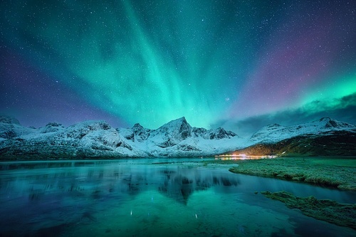 Northern lights over the snowy mountains, frozen sea, reflection in water at winter night in Lofoten, Norway. Aurora borealis and snowy rocks. Landscape with polar lights, starry sky and fjord. Nature