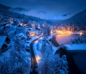 Aerial view of fairy town in snow, road, forest, Jasna lake and houses with lights at night in winter. Top view of mountain village, illumination, snowy pine trees at dusk in Kranjska Gora, Slovenia