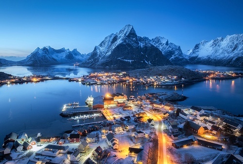 Aerial view of snowy village, islands, rorbu, city lights, blue sea, rocks and mountain at night in winter. Beautiful landscape with town, street illumination. Top view. Reine, Lofoten islands, Norway