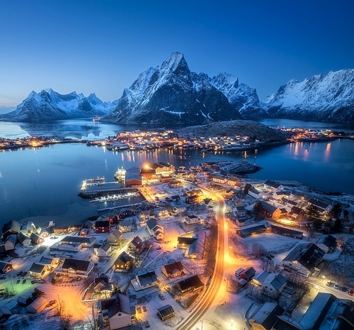 Aerial view of snowy village, islands, rorbu, city lights, blue sea, rocks and mountain at night in winter. Beautiful landscape with town, street illumination. Top view. Reine, Lofoten islands, Norway