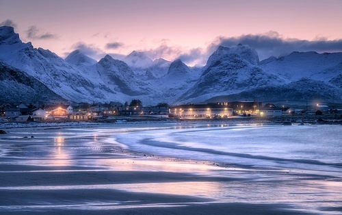 View of sandy beach, sea, waves, snowy mountains, street lights at winter night. Landscape with ocean, reflection, illumination, snowy rocks in clouds, pink sky at sunset. Lofoten Islands, Norway