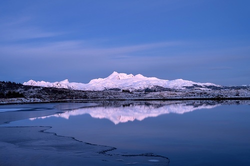 Snowy mountains, sea bay, reflection in water at sunset in winter. Lofoten islands, Norway. Colorful Landscape with rocks in snow, blue sky at dusk. Seashore at dusk. Nature background. Scenery