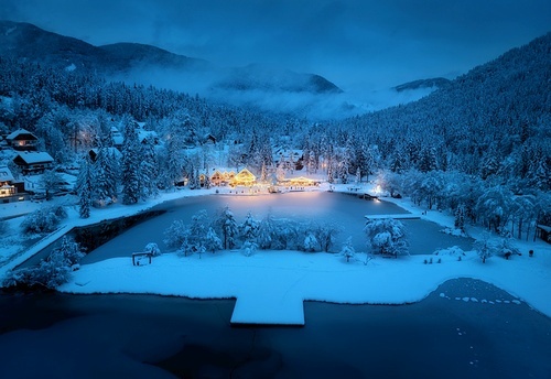 Aerial view of illuminated houses in fairy village in snow, forest, Jasna lake, street lights at winter night. Top view of alpine mountains in fog, snowy pine trees at dusk. Kranjska Gora, Slovenia