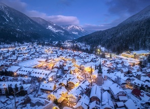 Aerial view of beautiful alpine village at cold winter night. Kranjska Gora, Slovenia. Top drone view of buildings in snow, street lights, road, snowy alpine mountains, sky at dusk. Triglav park