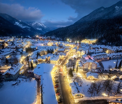 Aerial view of beautiful alpine village at cold winter night. Kranjska Gora, Slovenia. Top drone view of buildings in snow, street lights, road, snowy alpine mountains, sky at dusk. Triglav park