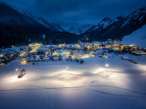 Aerial view of beautiful alpine village at cold winter night. Kranjska Gora, Slovenia. Top drone view of buildings in snow, street lights, road, snowy alpine mountains, sky at dusk. Triglav park