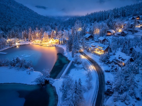 Aerial view of fairy alpine village in snow, road, forest, Jasna lake, houses, street lights at winter night. Top view of mountains, illumination, snowy pine trees at twilight. Kranjska Gora, Slovenia