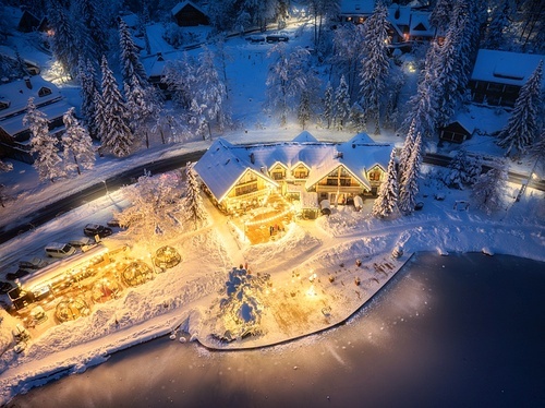 Aerial view of illuminated houses in fairy village in snow, forest, Jasna lake, street lights at winter night. Top view of alpine countryside, snowy pine trees at dusk. Kranjska Gora, Slovenia