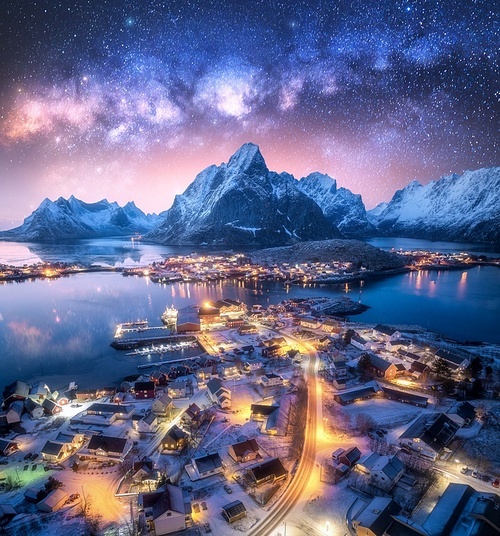 Milky Way over the snowy village, islands, rorbuer, city lights, sea, mountain, purple starry sky at winter night. Landscape with town, street illumination. Reine, Lofoten islands, Norway. Aerial view