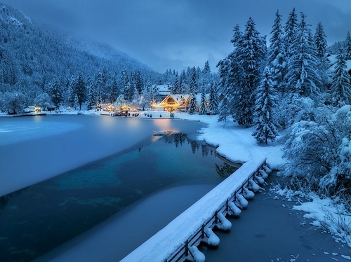 Aerial view of fairy alpine village in snow, bridge, forest, Jasna lake, houses, street lights at winter night. Top view of mountains, illumination, snowy pine trees at dusk. Kranjska Gora, Slovenia