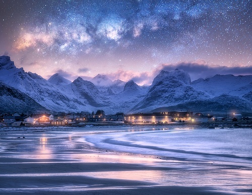 Milky Way over sandy beach, sea, waves, snowy mountains, street lights, starry sky at winter night. Landscape with ocean, reflection, illumination, snowy rocks, stars at dusk. Lofoten Islands, Norway