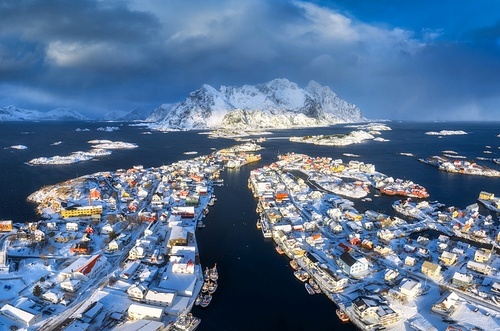 Aerial view of snowy Henningsvaer fishing village, Lofoten islands, Norway in winter at sunset. Top drone view of mountains in snow, sea, boats, bridge, road, cloudy sky, town, rorbuer and houses