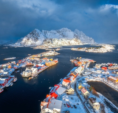 Aerial view of snowy Henningsvaer fishing village, Lofoten islands, Norway in winter at sunset. Top drone view of mountains in snow, sea, boats, bridge, road, cloudy sky, town, rorbuer and houses