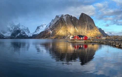 Red rorbuer on the island against snowy rocks lighted by sun, blue sea and dramatic cloudy sky in winter. Landscape with houses, mountains at sunset. Rorbu in Hamnoy village, Lofoten islands, Norway