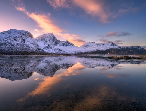 Beautiful snowy mountains and colorful sky with clouds at sunset in winter in Lofoten islands, Norway. Landscape with rocks in snow, sea coast, reflection in water at dusk, purple sky with pink clouds