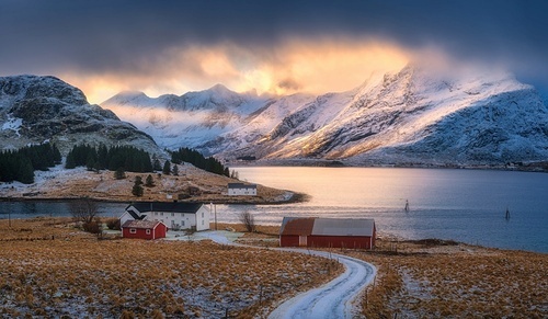 Rural road to the village near north sea and snowy mountains at sunset in winter. Landscape with red rorbu, field, sea, rocks in snow, golden sunbeams and orange low clouds in Lofoten Islands, Norway