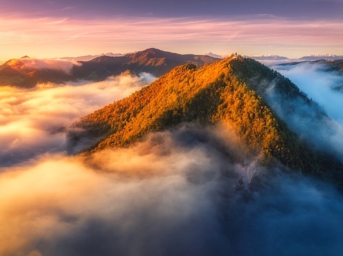 Aerial view of mountain peak in low clouds at sunrise in autumn. Top drone view of hills with red and orange trees in fog, colorful sky in fall. Slovenia. Nature. Mountain valley. Autumn forest. Alps