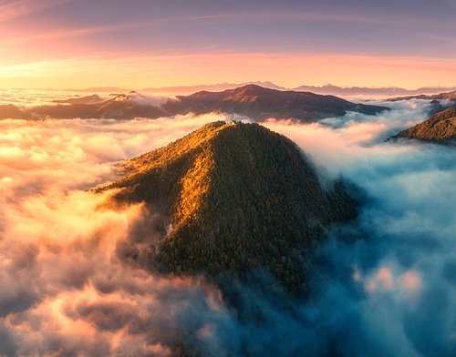 Aerial view of mountain peak in low clouds at sunrise in autumn. Top drone view of hills with red and orange trees in fog, colorful sky in fall. Slovenia. Nature. Mountain valley. Autumn forest. Alps