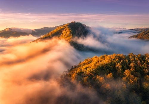 Aerial view of mountain peak in low clouds at sunrise in autumn. Top drone view of hills with red and orange trees in fog, colorful sky in fall. Slovenia. Nature. Mountain valley. Autumn forest. Alps