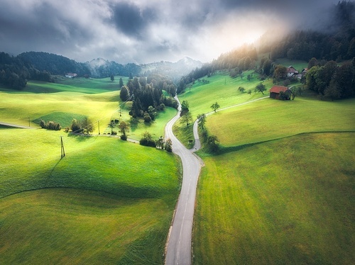 Aerial view of road in green meadows at sunset in summer. Top view from drone of rural road, mountains, forest. Beautiful landscape with roadway, sun rays, trees, hills, green grass, clouds. Slovenia