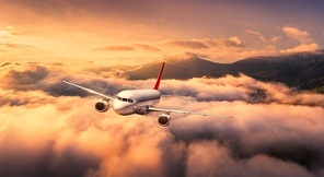 Plane is flying over mountain peaks in low clouds at golden sunrise. Top view of passenger airplane, colorful sky, hills in fog. Aircraft is taking off. Business travel. Commercial. Aerial view