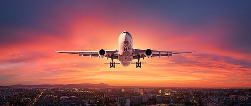 Airplane is flying in colorful sky over city at sunset. Landscape with passenger airplane, skyline, purple sky with red and pink clouds at dusk. Aircraft is landing at twilight. Aerial view of plane