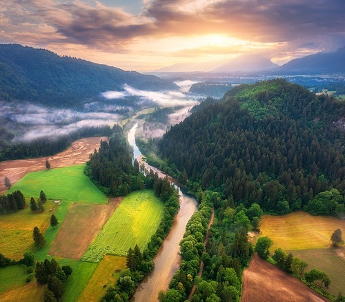 Aerial view of river in low clouds at sunrise in summer in Slovenia. Turns of river, green meadows and fields, grass and trees, rural road at golden dawn in spring. Top view of mountain countryside