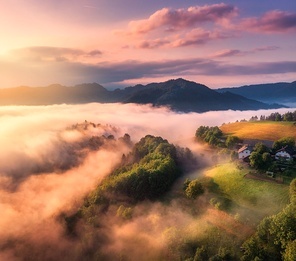 Aerial view of alpine meadows and mountains in low clouds at golden sunrise in summer. Top drone view of hills with green grass, trees in fog, house, colorful sky in Slovenia. Nature. Mountain valley