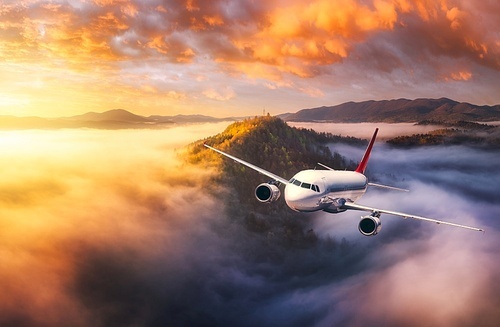 Plane is flying over mountain peaks in low clouds at golden sunrise. Top view of passenger airplane, colorful sky, hills in fog. Aircraft is taking off. Business travel. Commercial. Aerial view