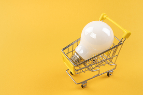 생활 속 에너지 - 노란색 배경지 위에 놓여진 불투명한 흰색 전구가 들어있는 미니어처 쇼핑 카트