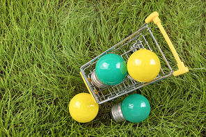 생활 속 에너지 - 푸른 잔디 위에 놓여진 노란색, 초록색 미니 전구들이 있는 미니어처 쇼핑 카트