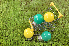 생활 속 에너지 - 푸른 잔디 위에 놓여진 노란색, 초록색 미니 전구들이 있는 미니어처 쇼핑 카트
