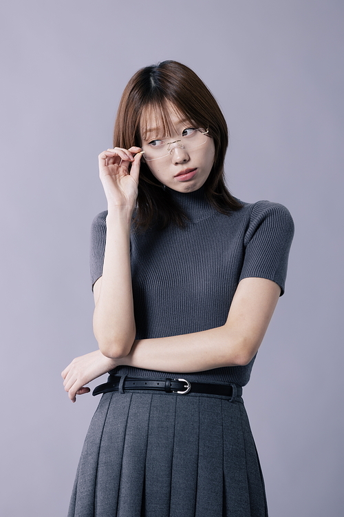 회색 의상을 입고 안경테를 만지며 포즈를 취하는 20대 한국인 여성