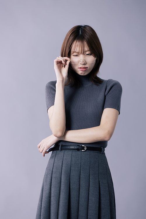 회색 의상을 입고 안경테를 만지며 얼굴을 찡그리는 20대 한국인 여성