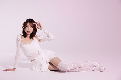 연핑크 배경의 흰색 발레코어룩을 입고 바닥에 앉아 포즈를 취하는 20대 한국인 여성