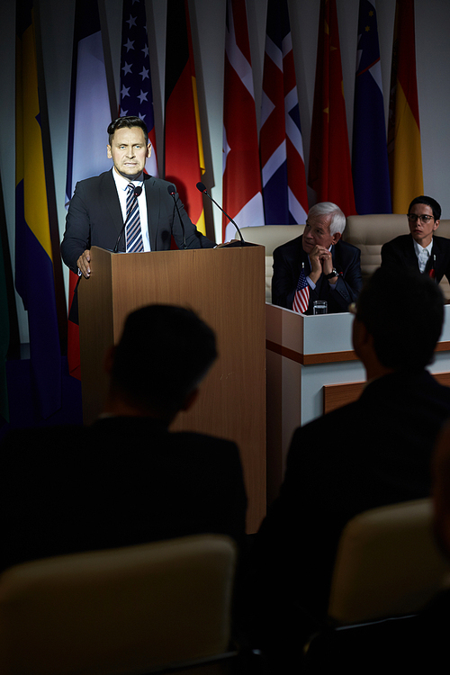 Formally-dressed concentrated speaker standing on stage, looking at the audience in conference room on background of national flags, ready for speech, face illuminated with light