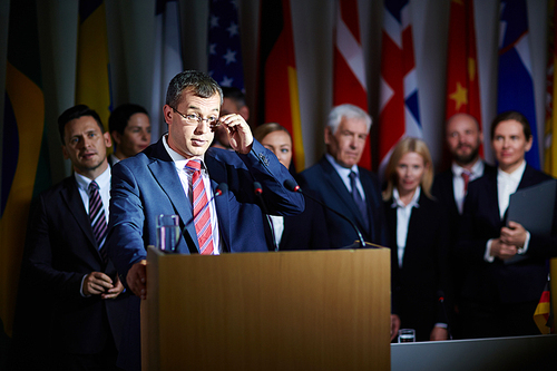 Formally-dressed spokesman standing at podium in conference hall, adjusting his glasses and making a pause in his speech, row of delegates and national flags behind him