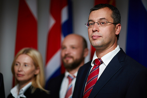 Close-up portrait of confident, attractive, middle-aged politician wearing glasses with his colleagues and national flags on blurred background
