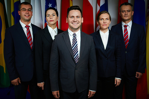 Group portrait of formal-dressed male and female delegates with confident smiling executive on foreground  on background of national flags