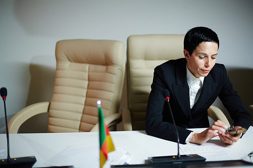 Attractive confident female delegate with short hair sitting on her break in conference room, using smartphone and smiling