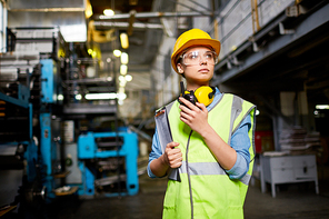 Young female in protective uniform, helmet and eyeglasses holding walkie-talkie while working at plant