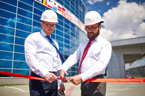 Happy architect cutting red ribbon while his co-worker holding it